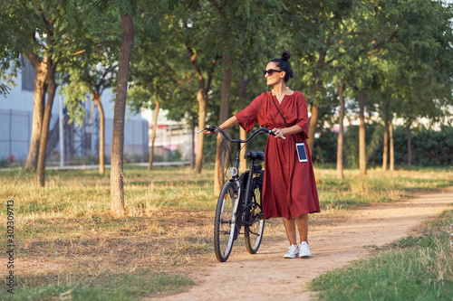 Full length lady in dress pushing bike and smiling while walking along path on sunny day in park
