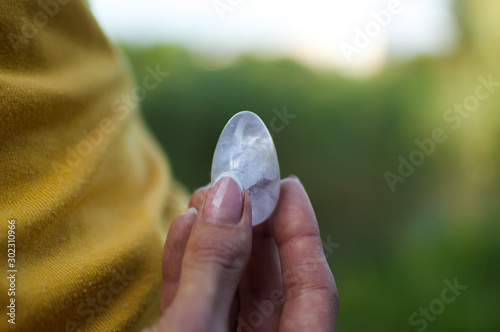 Adult girl in yellow shirt and blue jeans is holding transparent violet amethyst yoni egg for vumfit, imbuilding or meditation outdoors on her body background outdoors photo