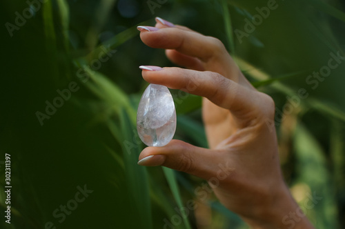 Female hand with french manicure holding transparent violet amethyst yoni egg for vumfit, imbuilding or meditation. Crystal quartz egg in hands on green background outdoors