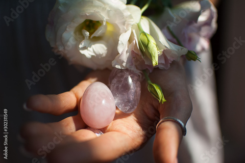 Female hands holding two yoni eggs for vumfit, imbuilding or meditation are made from pink quartz and transparent violet amethyst with white flowers indoors photo