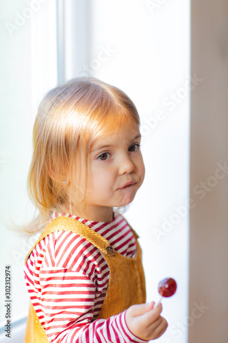 Portrait of cute toddler girl is sitting on windowstill with loly pop.