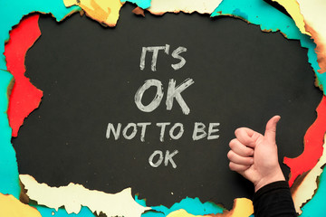 Text "It's OK not to be OK " in burned paper frame on black background and hand showing OK sign. Positive quote for persons suffering from personality disorder and Mental Health Awareness Month.