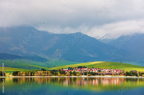 village on the shore of a lake. beautiful rural landscape in mountains. distant snow covered peaks in clouds. amazing scenery of western High Tatras in springtime