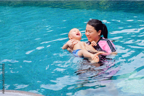 Smiling Asian mother & Cute little Asian 1 year old / 18 months toddler boy child relaxing playing in the swimming pool outdoor on nature, Mom and baby enjoying summer vacation at a tropical resort