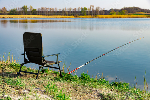 An empty chair and fishing rod on the lake coast in the morning. Green grass near large lake, blue sky, early hours