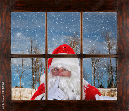 Santa Claus peeking in window making Shh sign, finger to mouth, with snowy winter scene in the background. photo