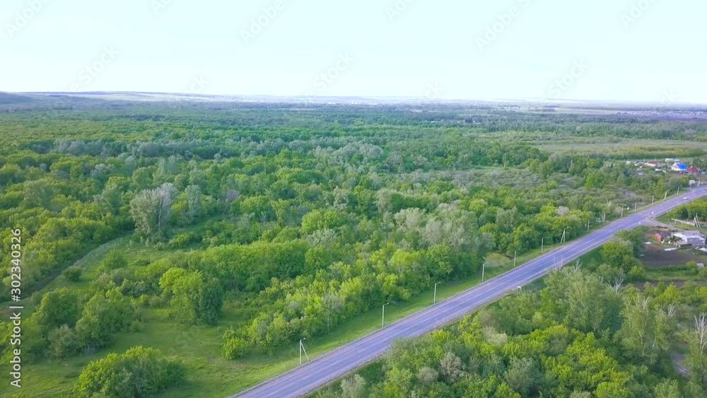 Aerial view of the road through the green forest to the small town on blue cloudy sky background. Art. Top view of fresh summer trees and the road.