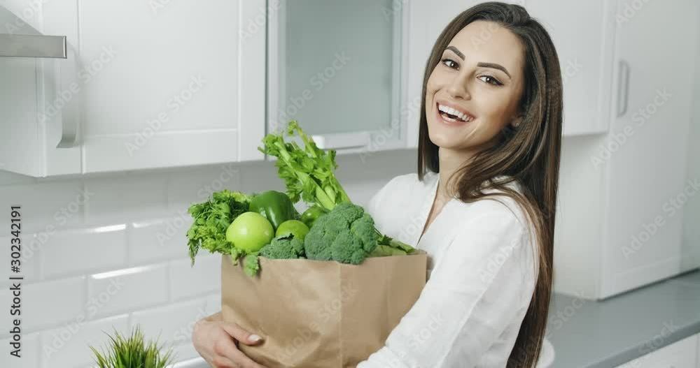 Attractive young woman holding bag with fresh green vegetables and smiling cheerfully, feeling fit and healthy Attractive young woman holding bag with fresh green vegetables and smiling cheerfully, feeling fit and healthy