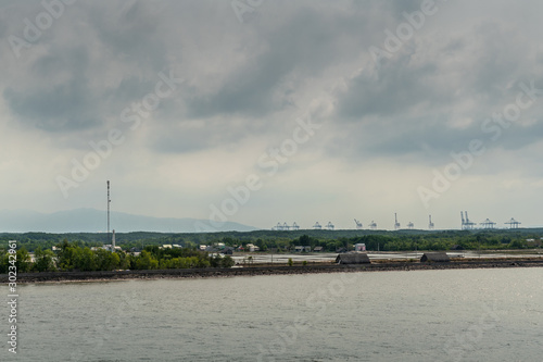 Long Tau River, Vietnam - March 12, 2019: At Thieng Lieng just up from mouth of river: International Container Terminal of Tan Cang visible behind water treatment plant under dark cloudscape. © Klodien