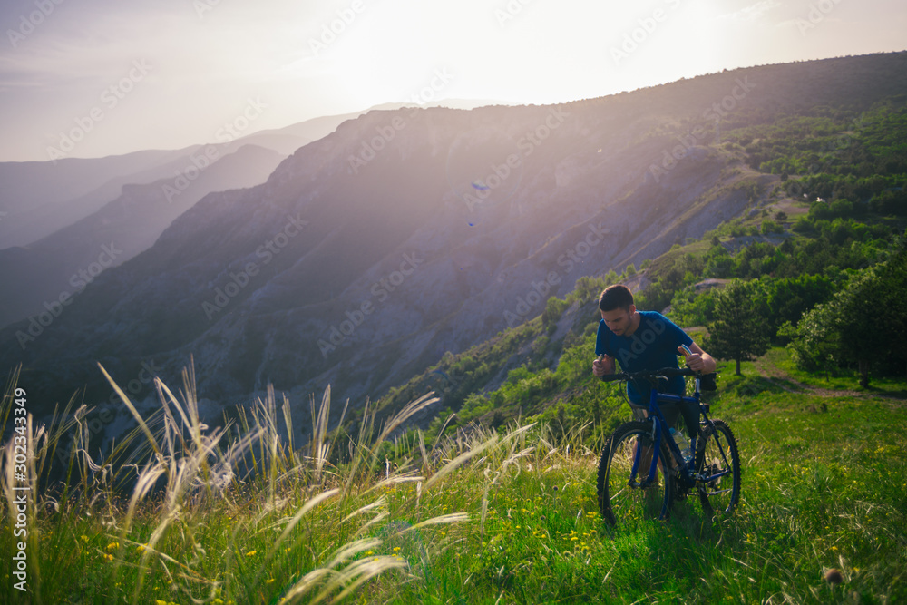 Perspective of a fit mountain biker pushing his bike uphill with amazing view on a forest, river and mountains in the background. Amazing green nature at sunset.