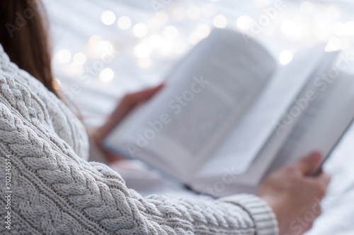 young woman wearing a beige sweater reading a book in a bed with white blanket and gold christmas lights photo