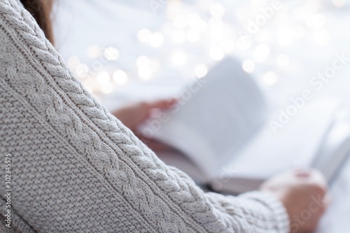 young woman wearing a beige sweater reading a book in a bed with white blanket and gold christmas lights photo