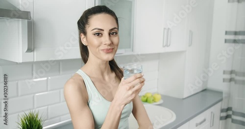 Attractive brunette drinking water after jogging and smiling to camera, keeping slim and fit, maintaining liquid balance
