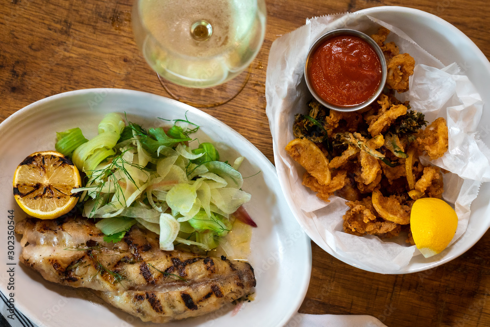 Grilled white fish with lemon and greens on a side and fried calamari rings with glass of white wine outside restaurant interior Grilled white fish with lemon and greens on a side and fried calamari rings with glass of white wine outside restaurant interior