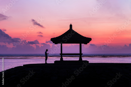 Outline of man standing next to seaside pagoda on Sanur beach, Bali, Indonesia at sunrise. Lavender sea in background, with clouds and multi colored sky. 