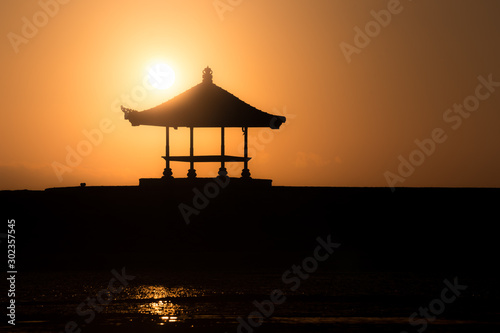 Cozy small traditional wooden house at Bali beach shot when sunset or sunrise. Enjoy fresh windy beach at small teak house in the morning or afternoon. Silhouette building with full round sun behind.