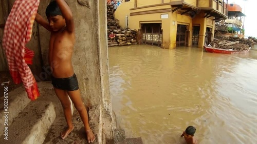 indian  boy wipes himself with chequered red material another boy go out from dirty water flood holy river Ganges piles of whirewood on boats and stairs Manikarnika Ghat place of cremation smashan flo
