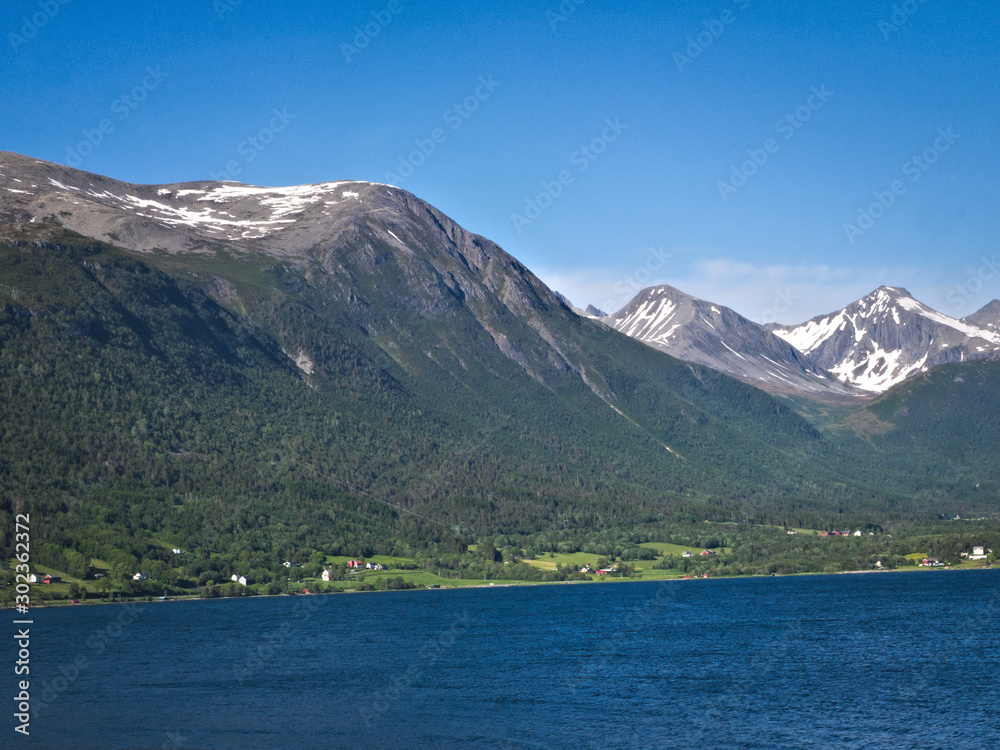 Isfjorden is the beautiful blue fjord at Andalsnes, Norway, with homes at the base of the mountains and snow and clear sky in the background on a sunny day.