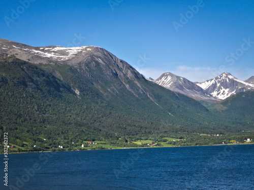 Isfjorden is the beautiful blue fjord at Andalsnes, Norway, with homes at the base of the mountains and snow and clear sky in the background on a sunny day.