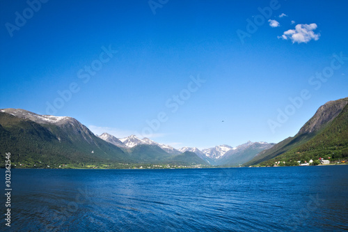 Isfjorden is the beautiful blue fjord at Andalsnes  Norway  with homes at the base of the mountains and snow and clear sky with a couple fluffy clouds in the background on a sunny day.