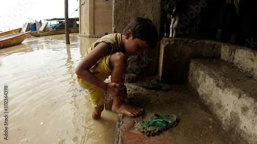 little indian boy washes his feet in dirty water of flooded Ganges beton stairs hoofs of cow boats on pier slippers in cow's shit Varanasi