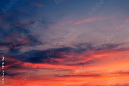 Light cirrus clouds at sunset are painted in bright fairy-tale fiery red, orange, golden, yellow colors against a dramatic dark blue sky. Abstract trendy modern texture background