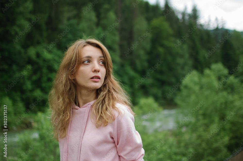 Amazing young girl in pink casual clothes with long hair on the hill with green forest background during spring or early autumn in the mountains.