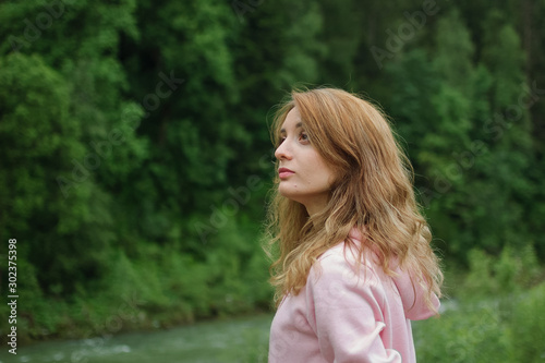 Side view of young calm blonde woman in pink clothes with long hair on the hill with green forest background during spring or early autumn in the mountains. Vacation or holiday concept