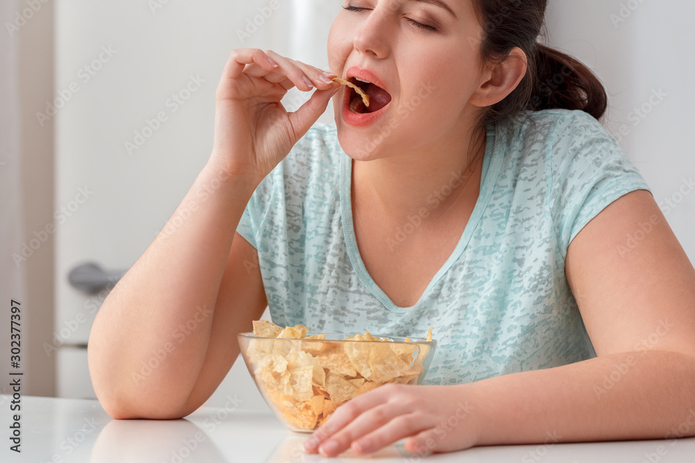 Breaking Diet. Chubby girl sitting at kitchen table eating chips closed eyes delightful close-up Breaking Diet. Chubby girl sitting at kitchen table eating chips closed eyes delightful close-up