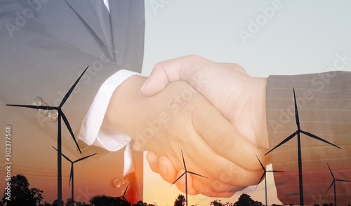Double exposure Businessman hand shake image of between sunset and wind energy turbine images.Electrical power engineering, environment and futuristic concept.