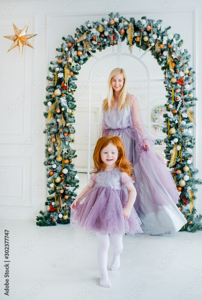 Redhead daughter and her blonde mother are playing before arch of fir branches in a Christmas studio