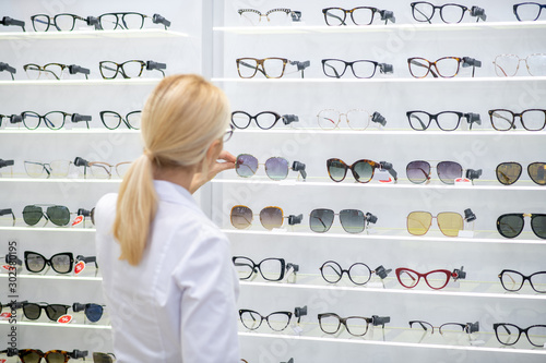 Blonde-haired ophthalmologist putting glasses on shelves in optical store