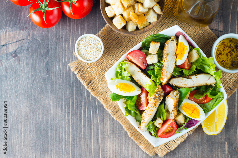 Fresh Caesar salad with delicious chicken breast, ruccola, spinach, cabbage, arugula, egg, parmesan and cherry tomato on wooden background. Oil, salt and pepper. Healthy and diet food concept.