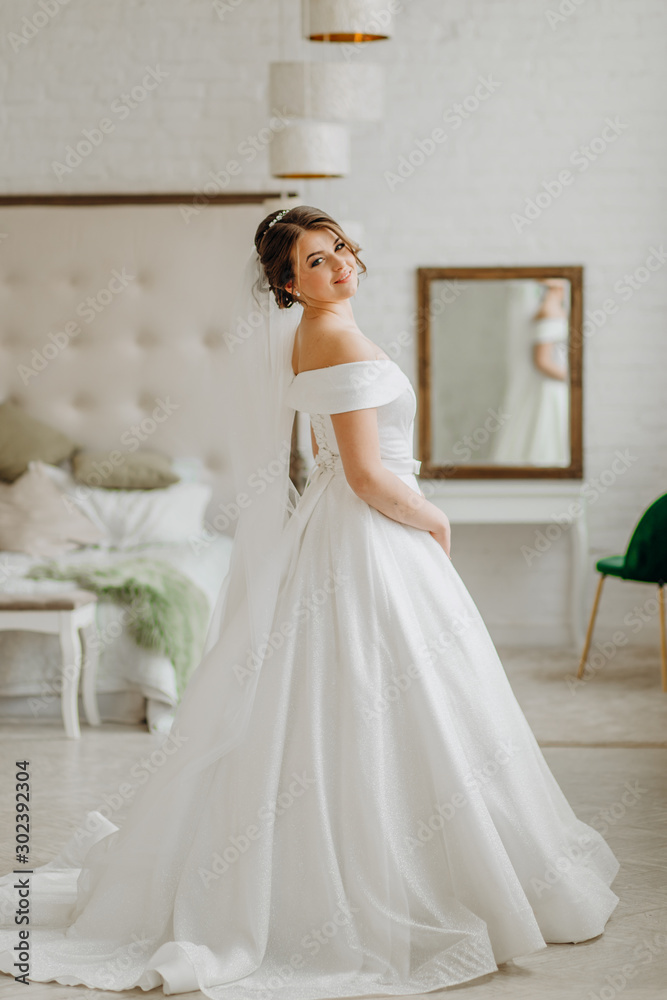 The bride in a white dress stands in a room by the bed