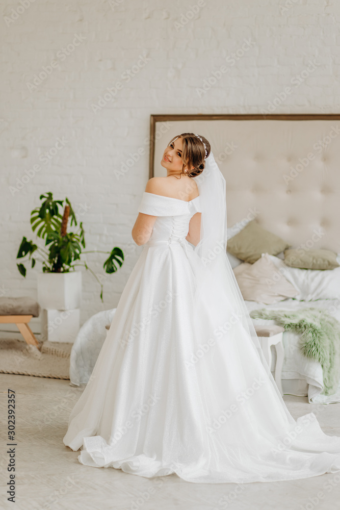 Dreamy bride in a white dress stands in a room by the bed Dreamy bride in a white dress stands in a room by the bed
