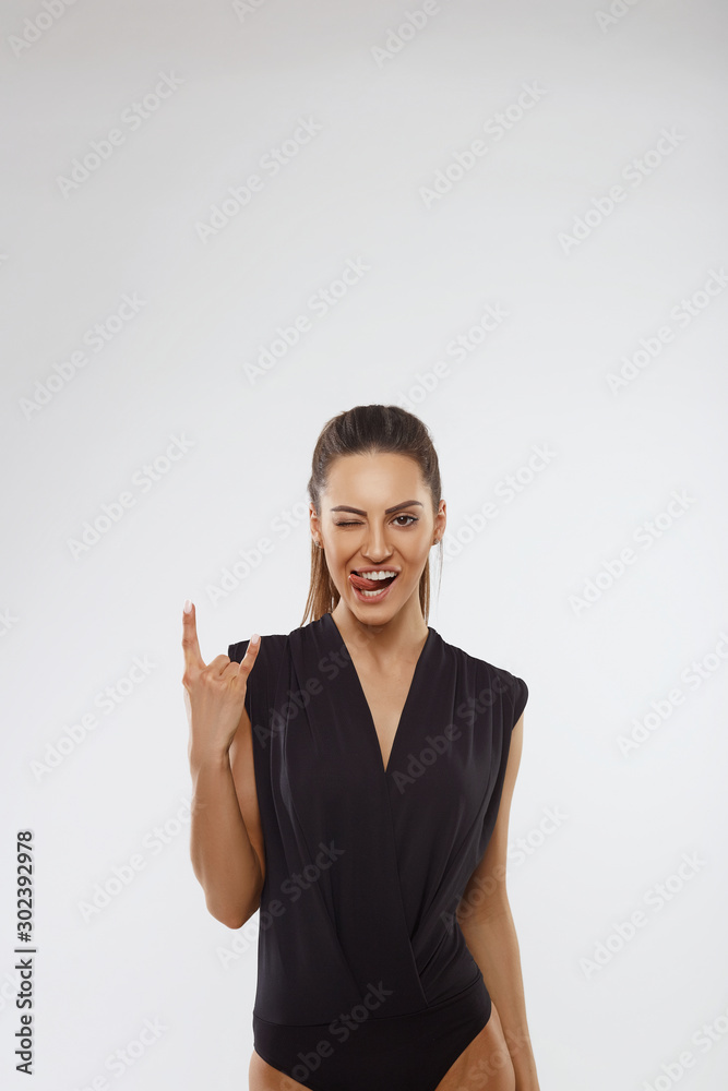 Crazy emotional young model showing the tongue and rock sign by hand on grey wall background. Closeup trendy portrait of woman Crazy emotional young model showing the tongue and rock sign by hand on grey wall background. Closeup trendy portrait of woman