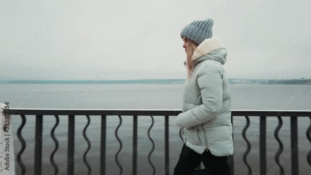 charming girl walks in a winter jacket and hat along the promenade and smiles