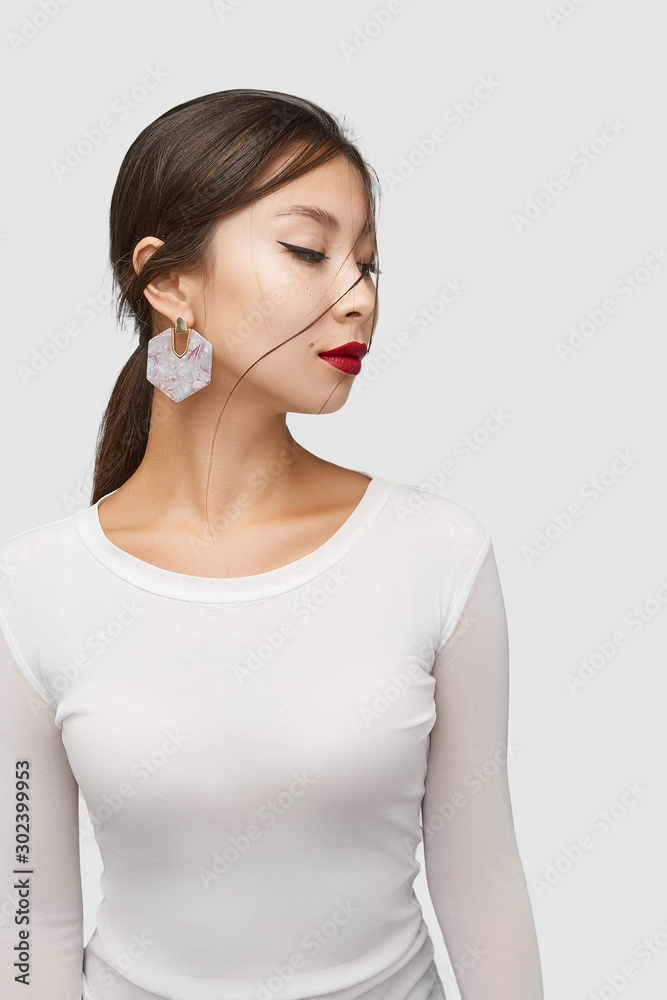 Medium shot of a young Asian woman in an ivory white blouse and with a golden stud earring made as a glimmering pearl gem plate in the form of a hexagon. Medium shot of a young Asian woman in an ivory white blouse and with a golden stud earring made as a glimmering pearl gem plate in the form of a hexagon.