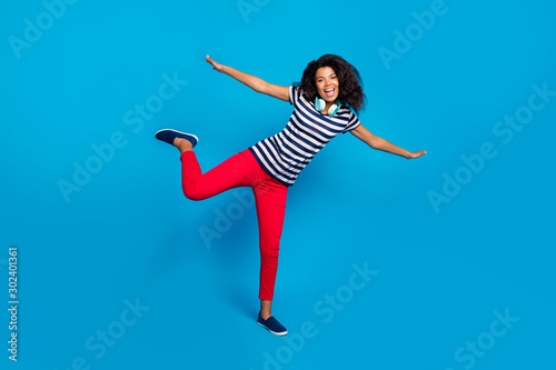Fototapeta Naklejka Na Ścianę i Meble -  Full length photo of positive funky funny afro american girl have rest headset to listen music hold hands imagine she fly like bird wear striped t-shirt casual outfit isolated blue color background