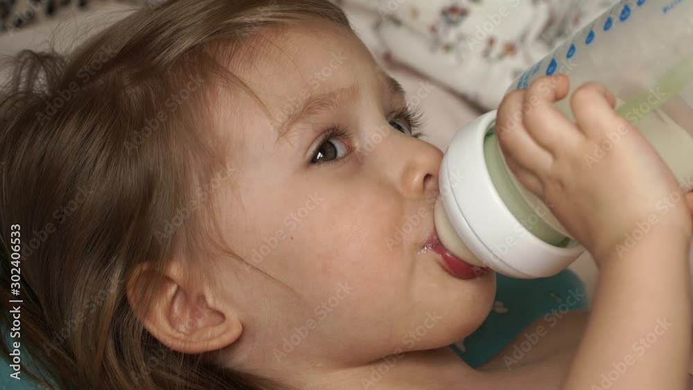 Сlose up of little cute baby girl lies on bed after sleep in morning and sucks milk from bottle with great appetite. Baby food. Infant formula milk concept