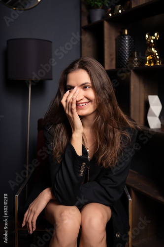 Smiling lady in mini skirt and oversized black jacket sitting on modern armchair in the room