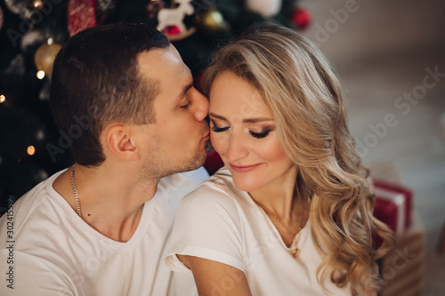 Headshot of beautiful young Caucasian couple against blurred Christmas tree. Boyfriend kissing his girlfriend in cheek with tenderness.