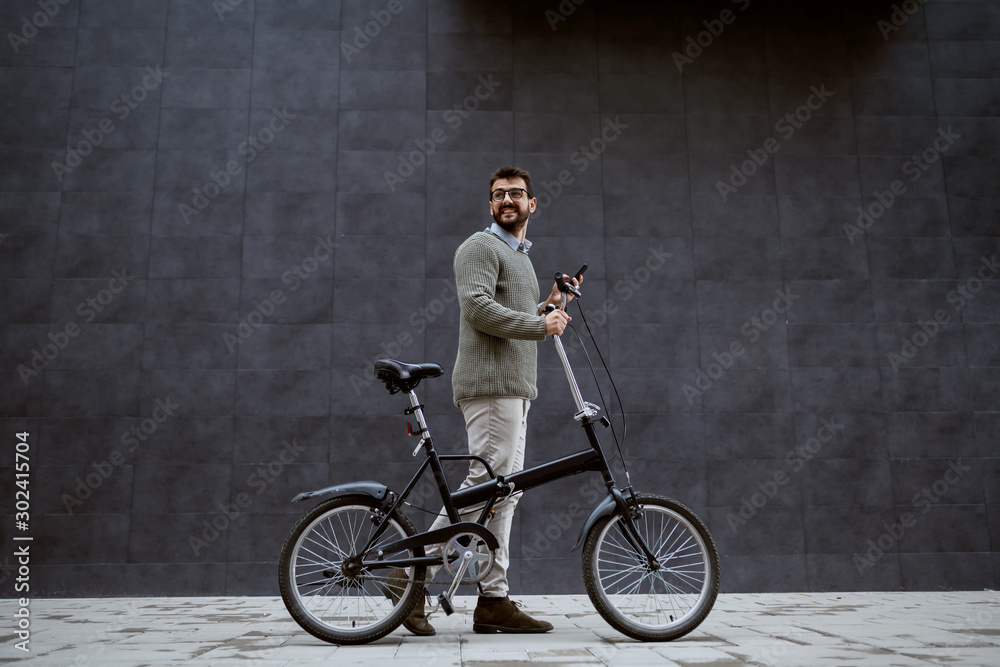 Full length of smiling caucasian fashionable man pushing bicycle and holding smart phone while passing by gray wall. Full length of smiling caucasian fashionable man pushing bicycle and holding smart phone while passing by gray wall.