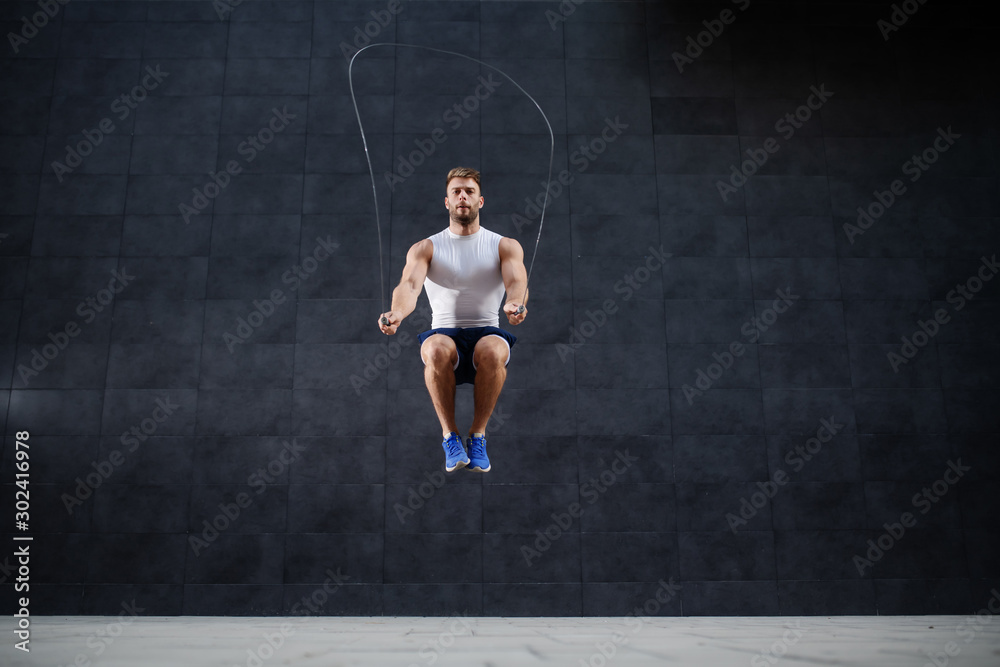 handsome muscular caucasian man in shorts and t-shirt skipping rope in front of gray wall outdoors.