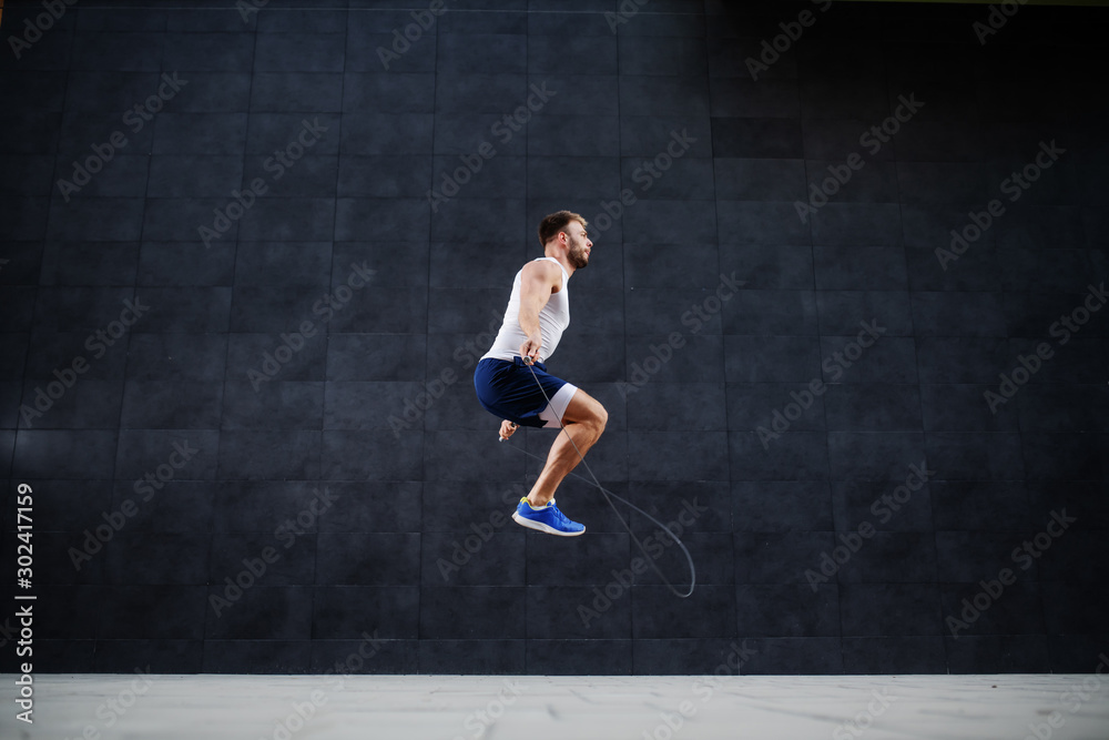 Side view of handsome muscular caucasian man in shorts and t-shirt skipping rope in front of gray wall outdoors.