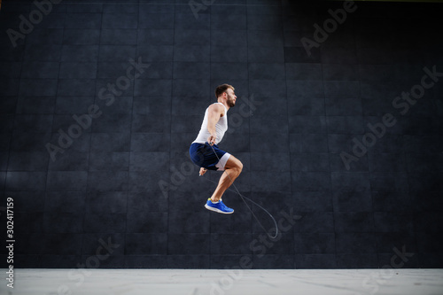 Side view of handsome muscular caucasian man in shorts and t-shirt skipping rope in front of gray wall outdoors.