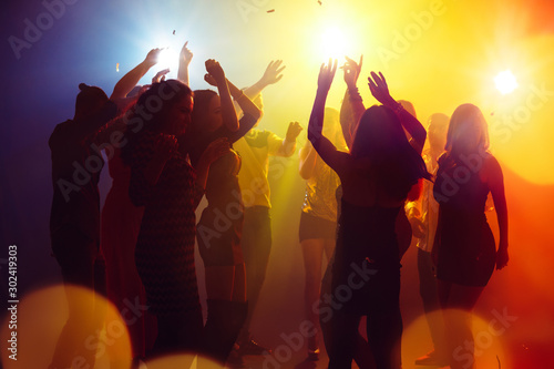 Holidays. A crowd of people in silhouette raises their hands on dancefloor on neon light background. Night life, club, music, dance, motion, youth. Yellow-blue colors and moving girls and boys.