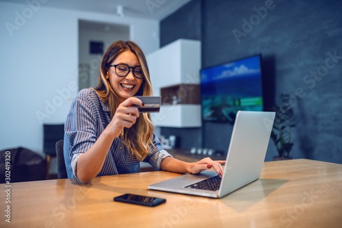 Young attractive smiling Caucasian woman with eyeglasses holding credit card and typing number of her bank account on laptop while sitting at dining table. Online shopping concept. photo