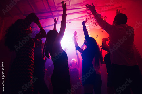 Catching moment. A crowd of people in silhouette raises their hands on dancefloor on neon light background. Night life, club, music, dance, motion, youth. Purple-pink colors and moving girls and boys.