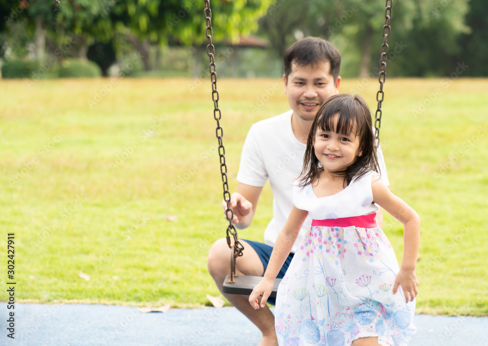 Asian father and daughter are playing the swing together in the playground of the park with happy moment, concept of outdoor activity for children in the family lifestyle.